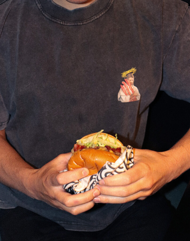 Person holding a gourmet burger wrapped in paper, wearing a T-shirt with a humorous bacon-themed graphic.